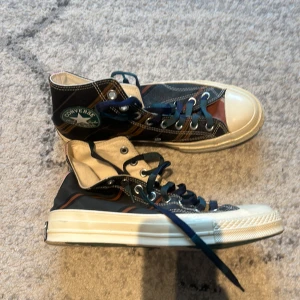 Converse Chuck Taylor Patchwork sneakers - Säljer ett par Converse Chuck Taylor sneakers i patchwork-stil med blå, bruna och svarta paneler. Skorna har klassisk vit tå, vita sulor och blå snörning. Converse-logga på sidan och cool retrovibe.