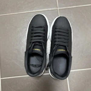 Svarta sneakers från Alexander McQueen - Säljer ett par svarta sneakers från Alexander McQueen med vit sula och svart ovandel. Skorna har rund tå, platt sula och klassisk snörning. Märkeslogga syns i guld på plösen och hälen.