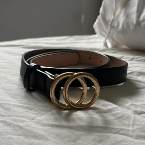 Svart bälte med dubbla guldringar - Stilrent svart bälte med två stora, runda guldfärgade ringar som spänne. Bältet har en slät yta och passar perfekt till jeans eller klänning för en trendig look.