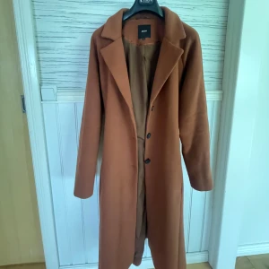 Brun kappa från Object - Säljer en snygg brun kappa från Object med klassisk krage och knäppning framtill. Kappan har bälte i midjan för en mer markerad siluett och långa ärmar. Perfekt för dig som vill ha en stilren och tidlös jacka till höst och vinter.