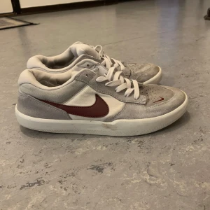 Nike SB Force 58 sneakers i grått och vitt - Nike SB Force 58 sneakers i ljusgrå mocka och vitt tyg med vinröd swoosh på sidan. Skorna har snörning, rund tå och platt sula. Perfekta för skate eller vardagsstil.