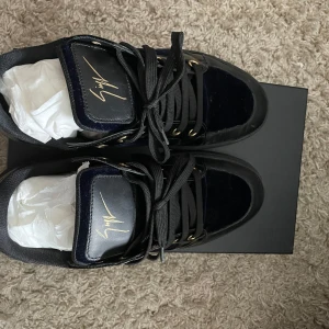  Giuseppe Zanotti - Säljer ett par svarta sneakers från Giuseppe Zanotti med guldfärgade detaljer och signatur på plösen. Skorna har svarta snören, platt sula och en mix av blankt och matt material. Snygg och lyxig design som sticker ut. Använd 5 eller 6 gånger. Box och dustbag ingår 