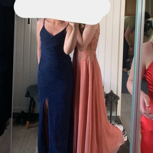 Mörkblå glittrig långklänning med slits - Säljer en elegant mörkblå långklänning  med glittrigt tyg, smala axelband och djup slits framtill. Klänningen har en figurnära passform och v-ringad urringning, perfekt för bal eller fest. Använd 1 gång