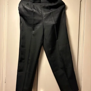 Svarta kostymbyxor med knäppning - Säljer ett par klassiska svarta kostymbyxor med pressveck och diskret glans. Byxorna har hög midja, raka ben och stängs med dragkedja och två metallknappar i midjan. Perfekta för en dressad look.