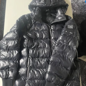  Peak Performance Dunjacka - Säljer en svart dunjacka från Peak Performance med huva och dragkedja. Jackan har en glansig finish och klassisk pufferstil. Märkeslogga på ärmen och praktiska fickor med dragkedja. Perfekt för kyliga dagar. Den har fått två små hål som knappt syns och går att så igen. 