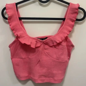 Rosa croppad linnetopp med volang - Supersöt rosa croppad linnetopp med breda axelband och volangdetalj längs ringningen. Toppen är ribbad och har en tight passform, perfekt för varma dagar.