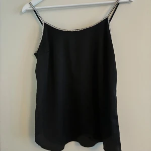 Svart linnetopp med strassband - Säljer en svart linnetopp med tunna axelband dekorerade med strass. Toppen har en slät och glansig yta som ger en elegant känsla. Perfekt för att lyfta en outfit med lite extra bling.