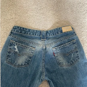 Blå bootcut jeans från Levi's - Säljer ett par klassiska blå jeans från Levi's med bootcut passform och låg midja. Jeansen har slitna detaljer på bakfickorna och en liten röd Levi's-tag. Perfekta för en avslappnad stil.
