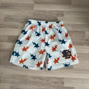 Snygga vita meshshorts från Eric Emanuel med blåa och orange stjärnor över hela tyget. Shortsen har resår i midjan och en broderad EE-logga på ena benet. Perfekta för sommaren och streetwear-stil.