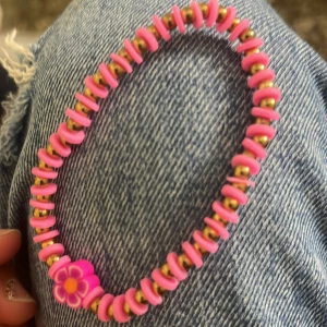 Rosa armband med blommor och gulddetaljer - Säljer ett färgglatt armband med rosa pärlor, små guldfärgade kulor och en söt blomdetalj i lila och gult. Perfekt accessoar för att liva upp din outfit!
