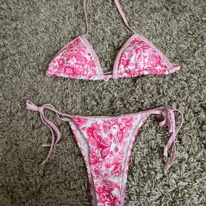 Rosa och vit blommig bikini med knyt - Säljer en snygg bikini i rosa och vitt med blommigt mönster. Överdelen är trekantsmodell med knyt i nacken och ryggen, och underdelen har höga ben och knyt i sidorna. Perfekt för sommaren!