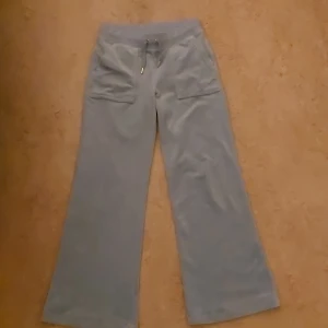 Ljusblåa velourbyxor från Juicy Couture - Säljer ett par ljusblåa velourbyxor från Juicy Couture med bred resår och snörning i midjan. Byxorna har raka, vida ben och fickor både fram och bak med knappdetaljer. Perfekta för en avslappnad och trendig look. De är i jätte bra skick har bara använt de bara 2 gånger