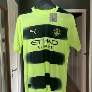 Manchester City bortatröja 2022/23 från Puma - Säljer en Manchester City bortatröja från Puma, säsongen 2022/23. Tröjan är neongul med svarta horisontella ränder, klubbmärke och Puma-logga på bröstet samt sponsortryck. Tillverkad i lätt och ventilerande material, perfekt för fotboll eller support. Helt ny tags är kvar 