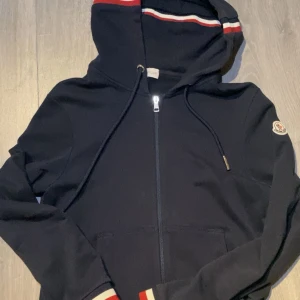Mörkblå hoodie från Moncler - Säljer en mörkblå hoodie från Moncler med dragkedja och huva. Tröjan har röda och vita detaljer på huva och ärmslut samt Moncler-logga på ärmen. Perfekt för en sportig och stilren look.