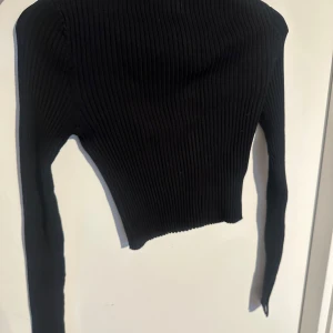 Svart ribbad croppad topp från H&M - Säljer en svart ribbad croppad topp från H&M. Toppen har lång ärm och är figurnära med stretchigt material. Perfekt att styla med höga jeans eller kjol. En kort del är öppen på båda armar vid handleden. 