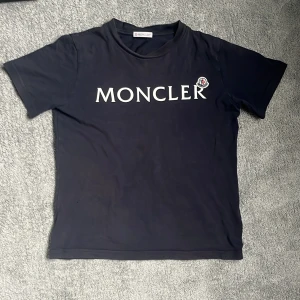 Svart t-shirt från Moncler med logga - Snygg svart t-shirt från Moncler med stor vit logga framtill och broderad patch på bröstet. Klassisk passform och rund halsringning. Perfekt för dig som gillar stilrena märkesplagg lite solblek 