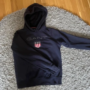 Mörkblå hoodie från GANT - Säljer en mörkblå hoodie från GANT med broderad logga och sköld på bröstet. Tröjan har huva och en klassisk känguruficka framtill. Perfekt för en avslappnad och stilren look.
