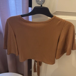 Brun ribbad croppad topp med knyt - Säljer en brun croppad topp med korta vida armar.  Bild 1 är framtill bild två är ryggen. 