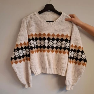 Vit stickad tröja med mönster - Säljer en vit stickad tröja med brunt och svart diamantmönster över bröstet och ärmarna. Perfekt för kyliga dagar!