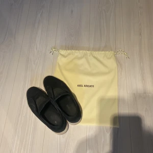 Axel Arigato laceless  - Säljer ett par stilrena laceless svarta sneakers från Axel Arigato med ovandel i mocka och vit platt sula. Skorna har en stilren design utan snörning och diskret logga på sidan. Perfekta för en clean och modern look. Påse ingår vid köp.