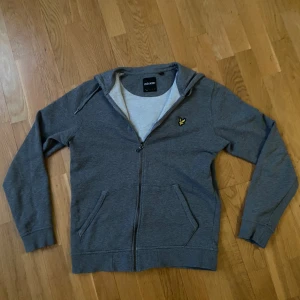 Lyle & Scott Hoodi - Säljer en grå hoodie från Lyle & Scott med dragkedja och huva. Tröjan är i storlek S och har två fickor framtill och klassisk gul logga på bröstet. Perfekt för en avslappnad stil och enkel att matcha med det mesta.