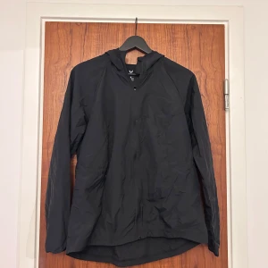 Svart löparjacka med huva från SOC - Svart vindjacka från SOC med huva och dragkedja framtill. Jackan är lätt, har lång ärm och är tillverkad i ett vindtätt syntetmaterial. Perfekt för blåsiga dagar och enkel att packa ner i väskan.