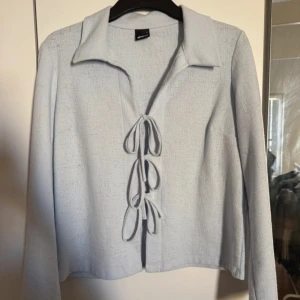 Ljusblå långärmad topp med knyt - Säljer en ljusblå långärmad topp från GinaTricot med krage och tre dekorativa knyt framtill. Tunnare material och rak passform, perfekt för lager-på-lager eller som statement-plagg.