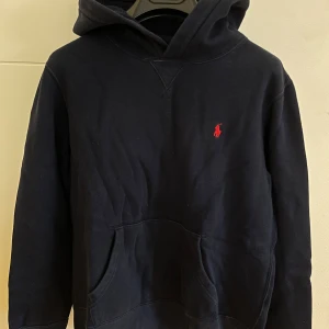 Mörkblå hoodie från Polo Ralph Lauren - Säljer en mörkblå hoodie från Polo Ralph Lauren med klassisk röd broderad logga på bröstet. Tröjan har huva, känguruficka och långa ärmar. Perfekt för en avslappnad och stilren look. Storlek: L (14-16). Hör av dig vid frågor!