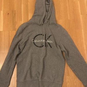 Grå hoodie från Calvin Klein Jeans - Säljer en grå hoodie från Calvin Klein Jeans med stor CK-logga på bröstet. Tröjan har huva med dragsko och långa ärmar. Perfekt för en avslappnad och stilren look.