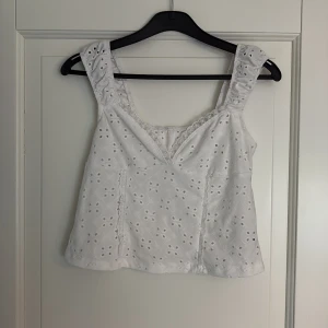 Vit broderad linnetopp med spetsdetalj - Säljer en söt vit linnetopp med breda axelband och v-ringning. Toppen har broderade blommor och en fin spetskant vid urringningen. Längd livet 26,5cm