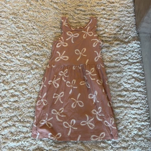 Rosa klänning med rosett-mönster från H&M - Supersöt rosa klänning från H&M med vita rosetter över hela tyget. Ärmlös modell med rund halsringning och utställd kjol. Perfekt för varma dagar och enkel att matcha med sneakers eller sandaler.