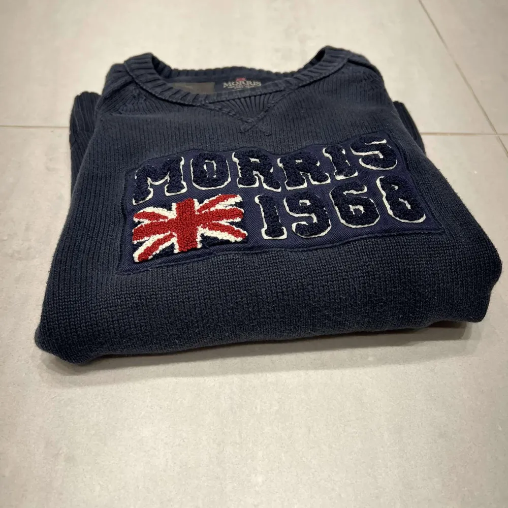 Mörkblå stickad tröja från Morris med broderad text 'Morris 1968' och brittisk flagga på bröstet. Tveka inte på att höra av er om mer bilder eller om ni har några funderingar 🤝. Neuletakit & Villapaidat.