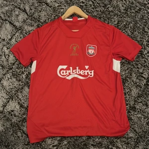 Liverpool FC European Champions 2005 fotbollströja - Röd Liverpool FC fotbollströja med Carlsberg-tryck, klubbmärke och broderad text 'European Champions Istanbul 2005' i guld. Kortärmad modell med vita detaljer på sidorna. Perfekt för fans av klassiska matcher.