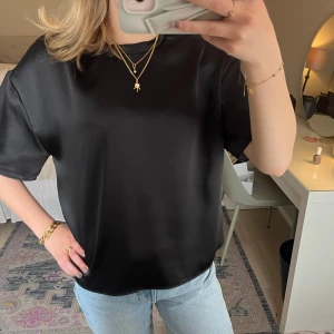 Svart satintopp - Säljer en svart, glansig satintopp med korta ärmar och rund halsringning. Toppen har en avslappnad passform och är perfekt att styla med jeans. Passar perfekt till en finare middag!!