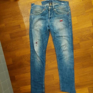 Blå jeans med feta hål och slitningar - Säljer ett par blå Dondup "George" jeans med slitningar och hål. Klassisk femficksmodell med knappgylf och raka ben. Jeansen har en tvättad look och detaljerade slitningar framtill.