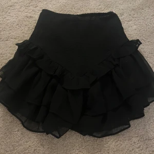 Svart volangkjol - Supersöt svart kortkjol med volanger och elastisk midja. Kjolen har flera lager volang som ger en riktigt snygg look. Perfekt att styla med en crop top eller stickad tröja.