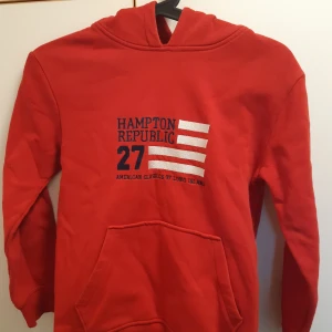 Röd hoodie från Hampton Republic - Säljer en röd (ser röd/orange ut på fotona men den är röd) hoodie från Hampton Republic med tryck framtill och stor ficka. Tröjan har huva och långa ärmar, perfekt för en avslappnad stil. Klassisk design med broderad logga och siffror på bröstet.