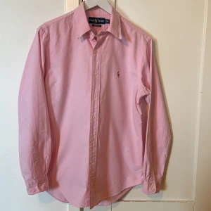 Rosa skjorta från Ralph Lauren - Klassisk rosa skjorta från Ralph Lauren med broderad logga på bröstet. Skjortan har lång ärm, knappar framtill och en normal passform (custom fit). Perfekt för en stilren look.