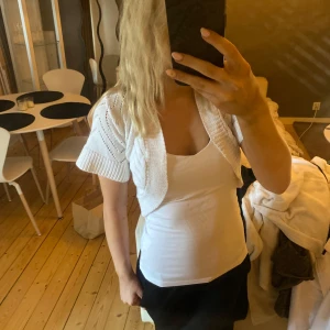 Vit stickad bolerotopp - Kort vit stickad bolerotopp med korta ärmar och öppen front. Perfekt att slänga över ett linne eller klänning för en extra touch. Snyggt mönster i stickningen ger en trendig look.