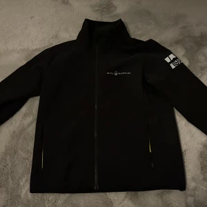 Svart jacka från Sail Racing - Svart jacka från Sail Racing med hög krage och hel dragkedja framtill. Jackan har två fickor med dragkedja och reflekterande detaljer på ärmar och rygg. Logotyp på bröstet och ärmen. Perfekt för en sportig stil.
