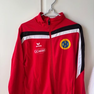 Röd träningsjacka från Erima med klubbmärke - Röd träningsjacka från Erima med dragkedja och klubbmärke på bröstet. Jackan har svarta och vita detaljer på axlarna och är långärmad. Perfekt för fotbollsträning eller uppvärmning.