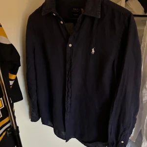 Mörkblå linneskjorta från Polo Ralph Lauren - Säljer en klassisk mörkblå skjorta i linne från Polo Ralph Lauren. Skjortan har lång ärm, knappar framtill och den ikoniska broderade loggan på bröstet. Perfekt för dig som gillar en stilren och tidlös look.