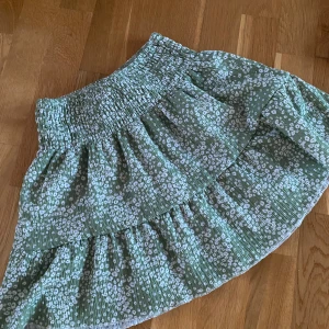 Grön blommig volangkjol - Söt grön kjol med vitt blommönster och volangdetaljer. Kjolen har en bred, smockad midja som ger en bekväm passform och ett luftigt fall. Perfekt för varma dagar och enkel att matcha med olika toppar.