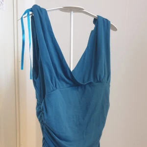 Blå omlottklänning från Seductions - Snygg blå omlottklänning från Seductions med djup v-ringning och knytband på axeln. Klänningen har draperad effekt längs sidorna och är tillverkad i ett mjukt material som känns bekvämt mot huden.