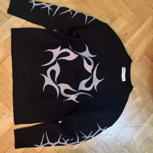 Svart stickad tröja med tribal-mönster - Svart stickad tröja från Bershka i storlek M med grått tribal-inspirerat mönster på bröstet och längs ärmarna. Tröjan har rund halsringning och långa ärmar. Perfekt för dig som gillar edgy och unik stil.