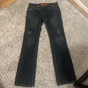 Mörkblåa jeans - Säljer ett par mörkblå jeans med klassiska kontrastsömmar och broderad logga på bakfickan. Jeansen här bootcut, slitna detaljer framtill och coola läderdetaljer vid midjan. Lite sönder längst midjan men inget som försämrar funktionen. För små för mig så finns ingen bild på