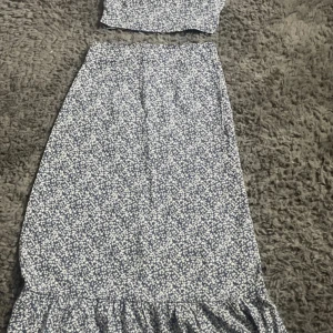 Blommig blå kjol och topp  - Tvådelad kjol och topp i blått med små vita blommor. Toppen har kort puffärm och rynkade detaljer, kjolen är lång med volangkant nertill och dragkedja på baksidan. Perfekt för sommarens alla tillfällen och ger en romantisk vibe.