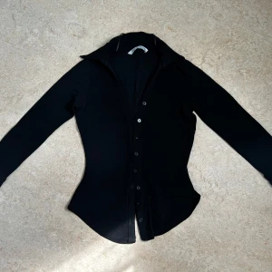 Svart skjorta från Zara  - Snygg svart skjorta från Zara med lång ärm och klassisk krage. Skjortan har en figurnära passform och knäppning framtill med svarta knappar. Perfekt att styla till jeans eller kjol för en clean look. Perfekt för jobbet