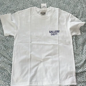 Vit t-shirt från Gallery Dept. - Vit t-shirt från Gallery Dept. med tryck i mörkblått framtill och stort tryck på ryggen. Klassisk passform, rund hals och korta ärmar. Tillverkad i mjuk bomull, perfekt för en clean och stilren look.