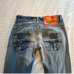 Blå jeans från Replay med broderad detalj, low waist  - Snygga blå jeans från Replay med en unik broderad detalj på baksidan. De är bootcut med slitna detaljer på benen. Midjemått: 39 cm, Innerbenslängd: 80 cm, Grenmått: 17cm